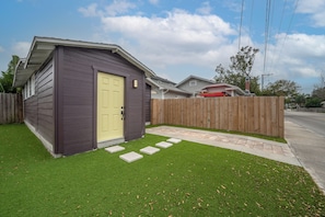 A small wooden shed with a yellow door, a wooden fence, and a paved pathway leading to it.