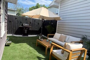 A backyard with a wooden fence, a barbecue grill, a wooden table, and a white sofa with cushions.