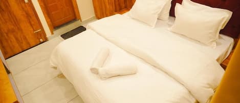 A hotel room with a double bed, two pillows, a rolled towel, wooden flooring, and a wooden door.