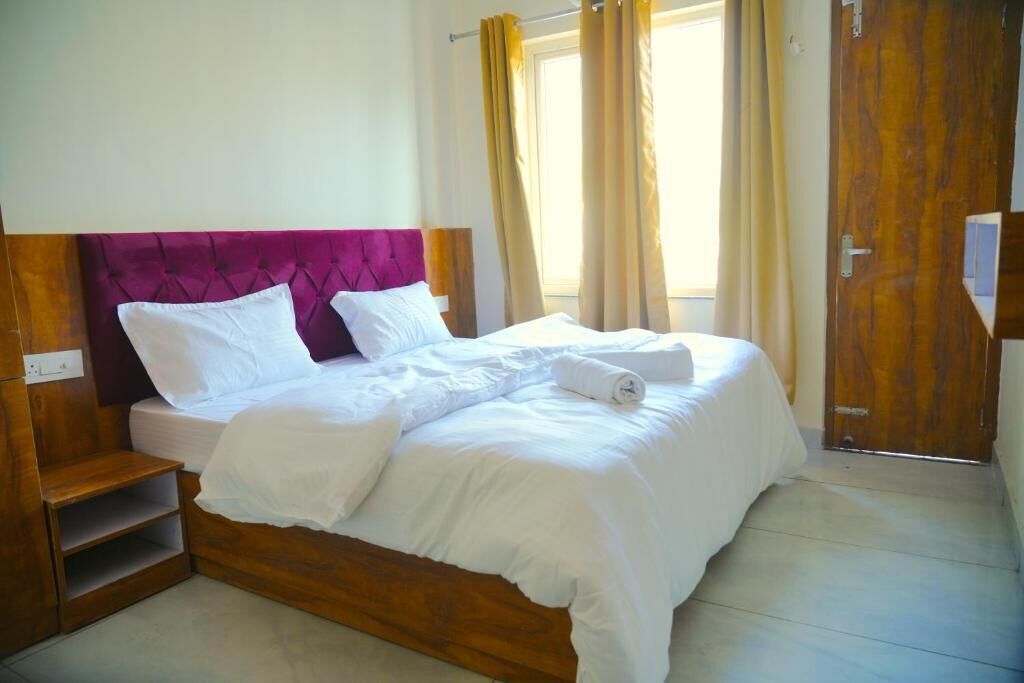 A double bed with a wooden frame and a purple headboard, accompanied by a wooden nightstand and a small shelf.
