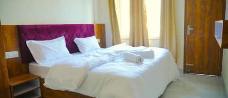 A double bed with a wooden frame and a purple headboard, accompanied by a wooden nightstand and a small shelf.