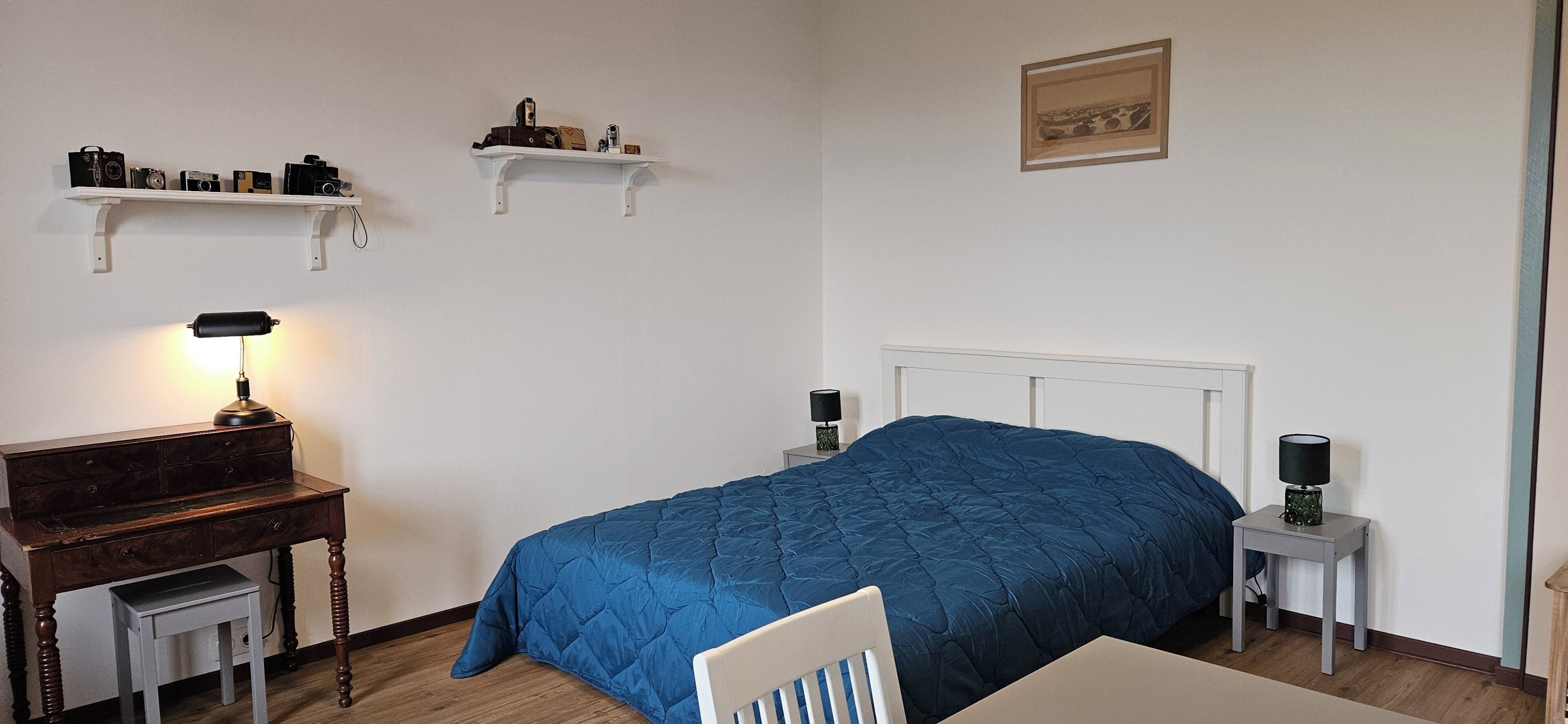 Et soveværelse med en seng, et skrivebord, en stol, en lampe og to vægmonterede hylder med forskellige genstande.