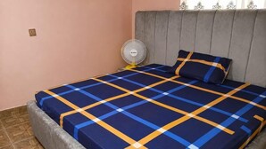 Un llit amb un estenat de quadres, un ventilador al costat del llit i una finestra amb cortines.