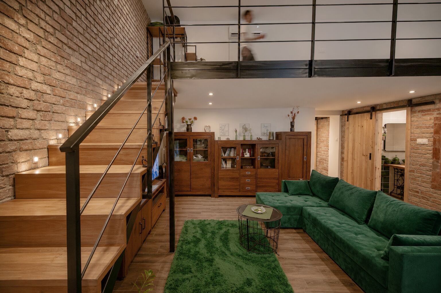 Een moderne woonkamer met een trap, een groene bank, houten kasten en een metselwerk muur.