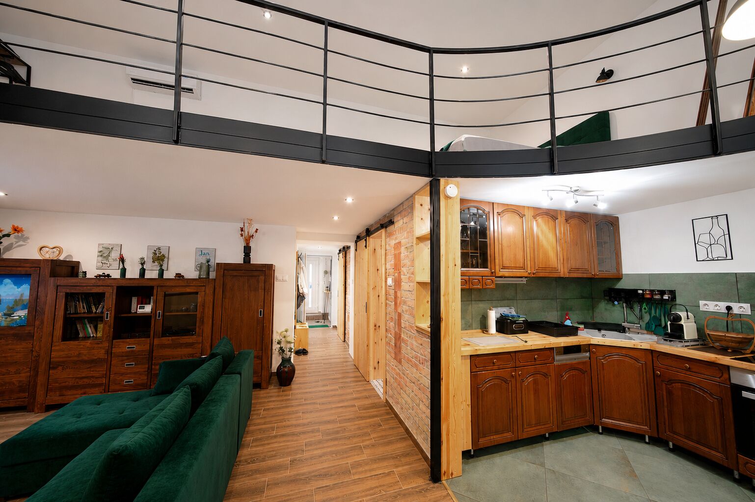 A modern kitchen with wooden cabinets, a green sofa, and a loft bedroom with a black railing.