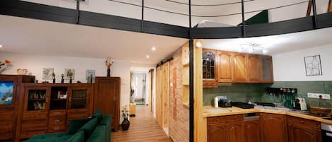 A modern kitchen with wooden cabinets, a green sofa, and a loft bedroom with a black railing.