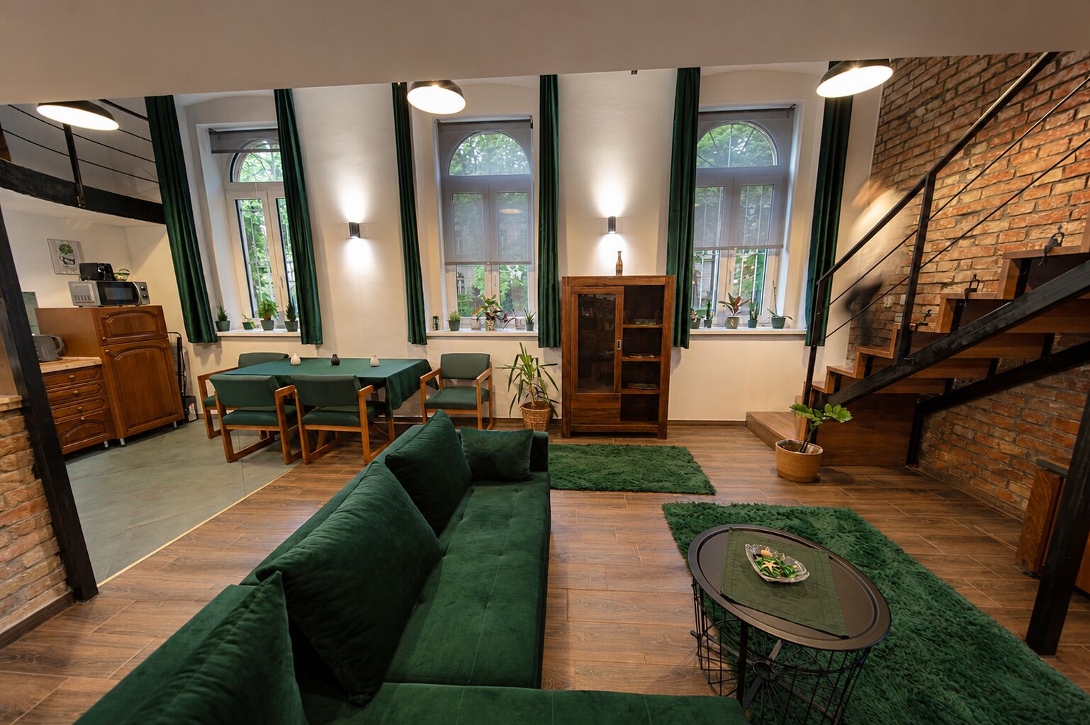A modern living room with a green sofa, dining table, and chairs, a staircase, and large windows with curtains.