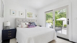 A bedroom with a large bed, a nightstand, a lamp, and a view of the outdoors through a sliding glass door. - Completely Renovated Sag Harbor Cottage with Lush Gardens, Pergola, and... (Sag Harbor)