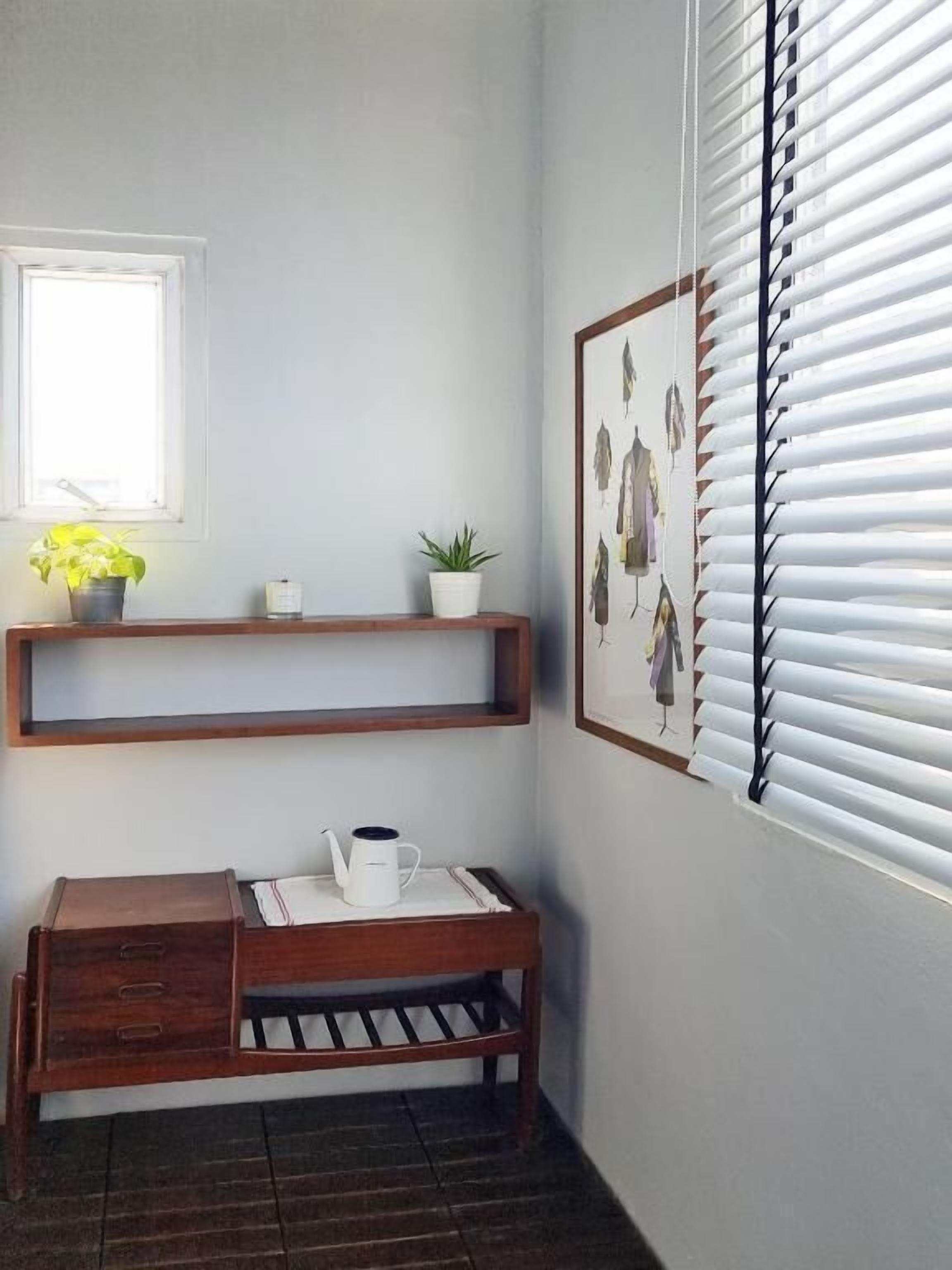 A corner room with a wooden bench, a small table with a white mug, a window with blinds, and a wall-mounted shelf with plants.