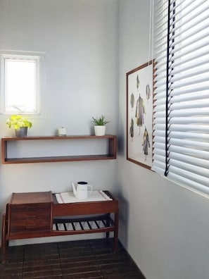 Habitación en esquina con un banco de madera, una mesita con una taza blanca, una ventana con persianas y un estante con plantas.