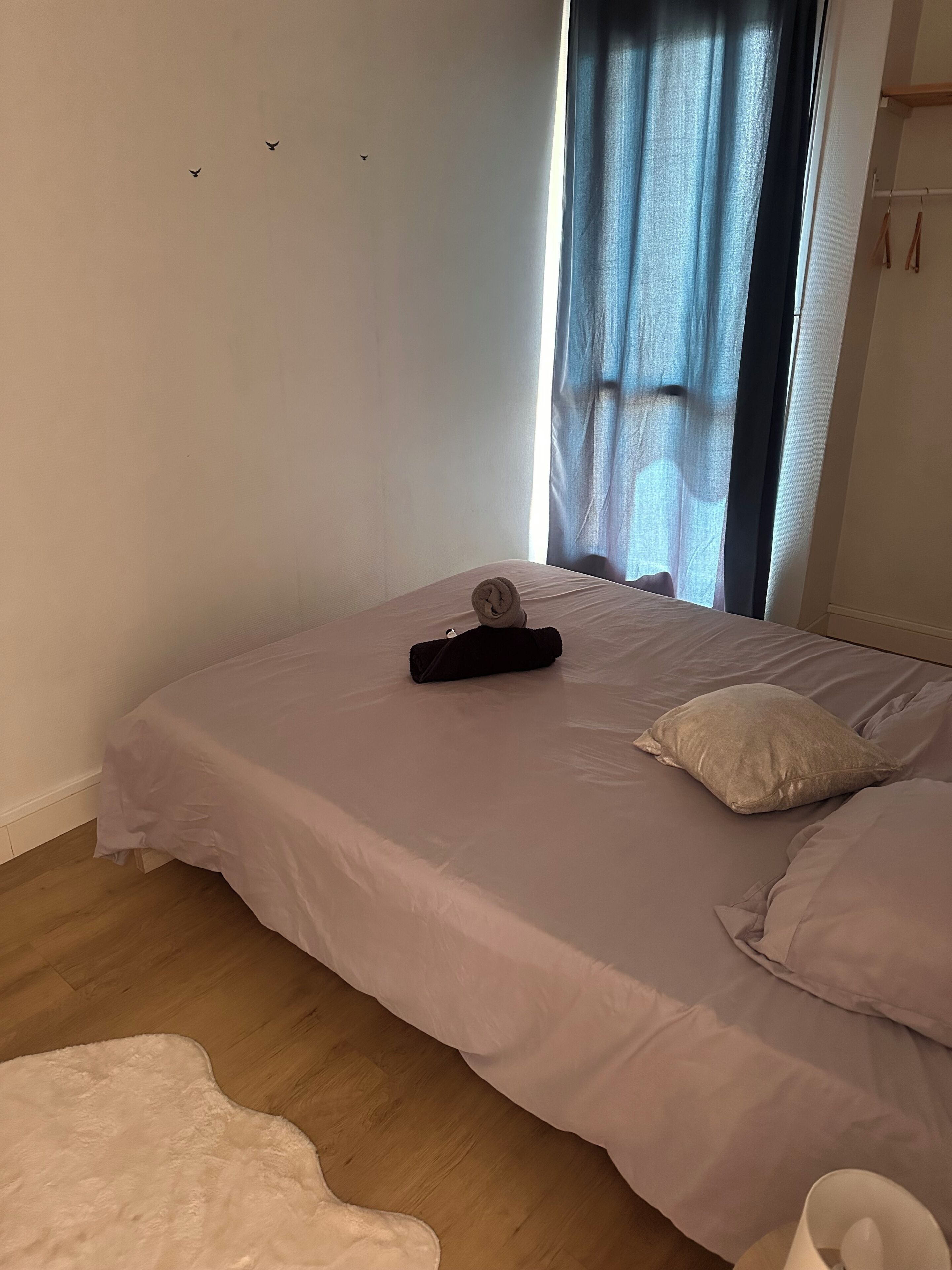 Una cama bien tendida con una toalla enrollada y un almohadón, una ventana con cortinas y un piso de madera.