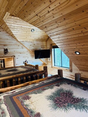 A room with a sloped ceiling, wooden walls, a large window, a flat-screen TV, and a patterned rug. - Deer Creek Lodge -12 min. to YNP+Hot tub+AC+Sauna Sleeps 26 + New Built 2026 (Island Park)