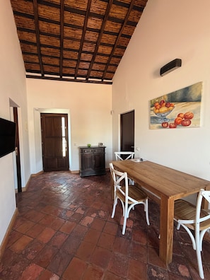 Un coin repas avec une table en bois et des chaises blanches, un sol carrelé, un buffet en bois foncé et un tableau accroché au mur.