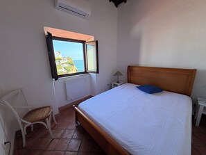 Ein Schlafzimmer mit Bett, Sessel, Nachttisch, Lampe und Fenster mit Blick auf eine Küstenlandschaft.