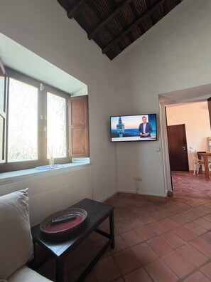 A living room with a television mounted on the wall, a window with wooden shutters, a small table with a remote control, and a door leading to another room. - A House by the Atlantic Ocean in Carvoeiro (Carvoeiro)