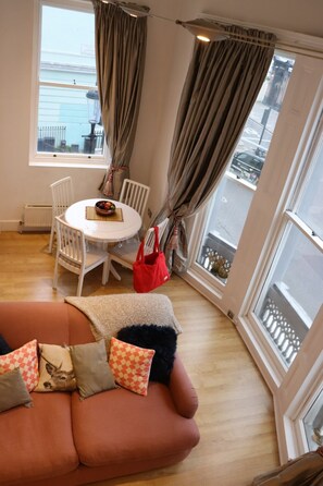 A cozy living room with a sofa, a dining table with chairs, a red bag, and large windows with curtains. - Mezzanine studio flat in the heart of Notting Hill (London)
