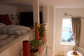 A bedroom with a bed, a TV, a window with curtains, a potted plant, and a small table with a bowl. - Mezzanine studio flat in the heart of Notting Hill (London)
