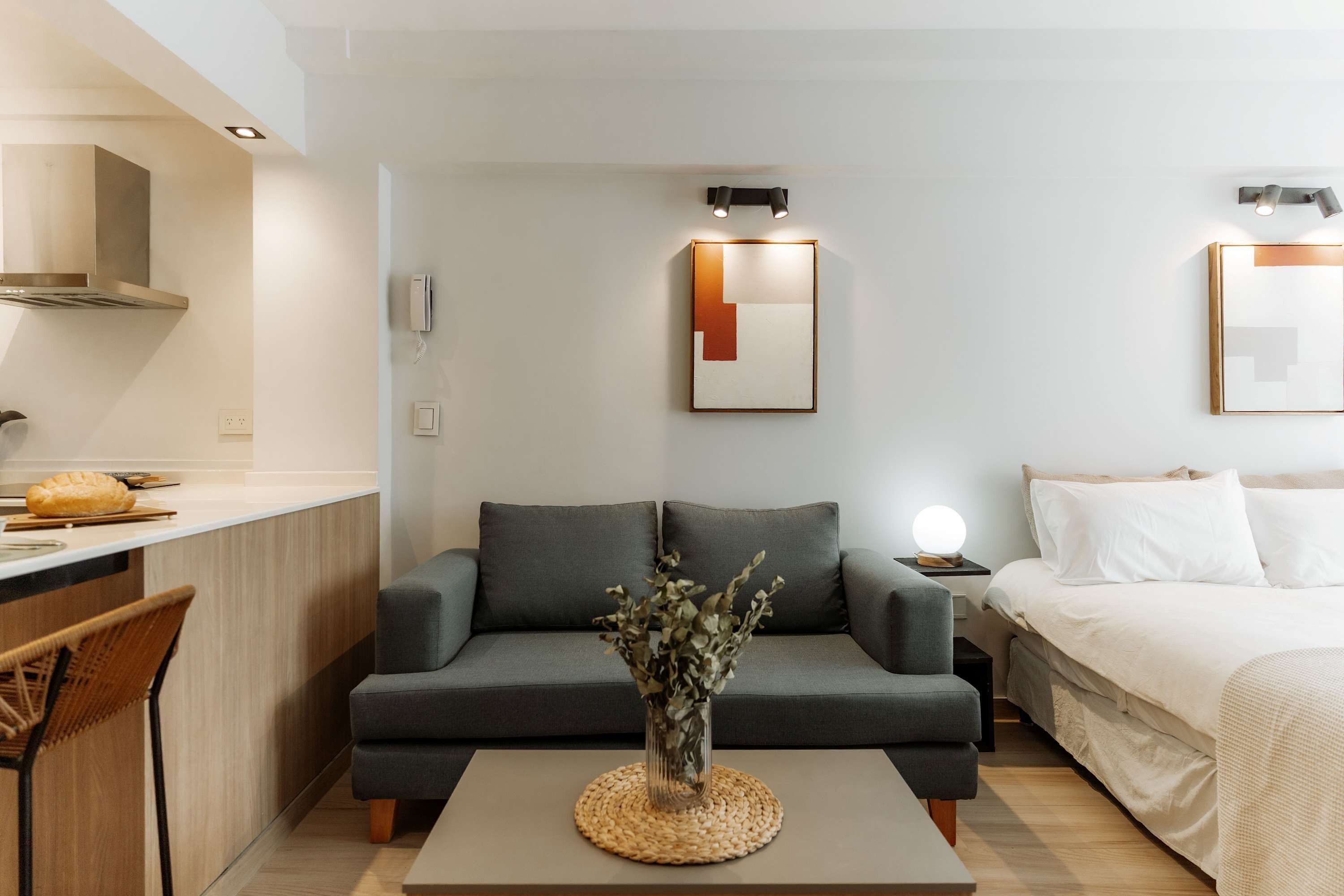 Una habitación de hotel moderna con sofá, mesita de noche con lámpara, cama, una pequeña mesa con un florero y una zona de kitchenette.