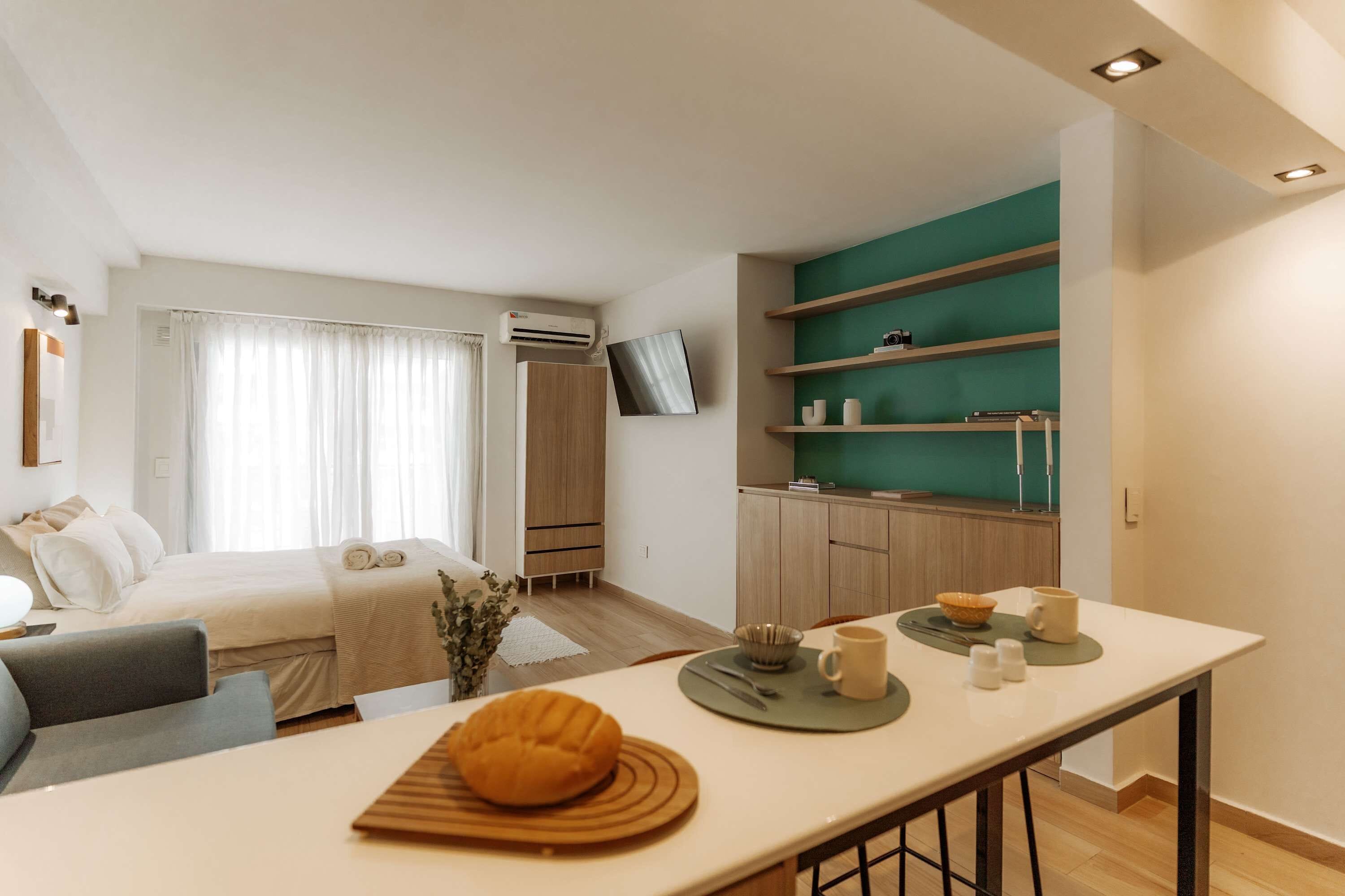 A modern hotel room with a bed, a desk with a chair, a kitchenette, and a TV mounted on the wall.