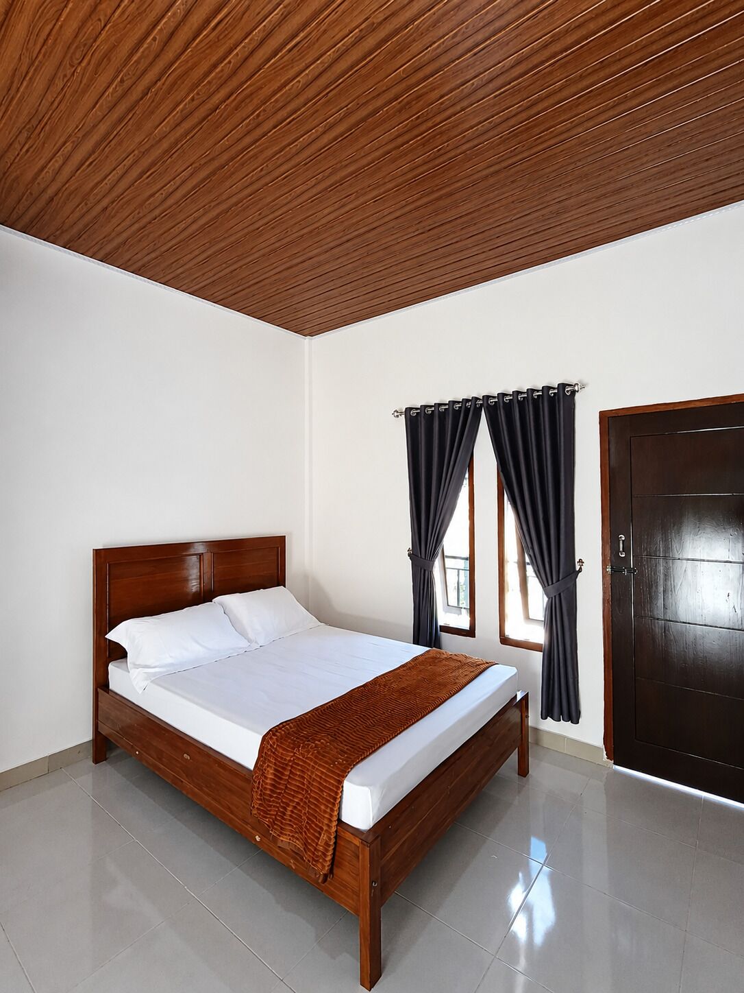 A bedroom with a wooden bed, white bedding, a brown throw blanket, a window with curtains, and a dark door.