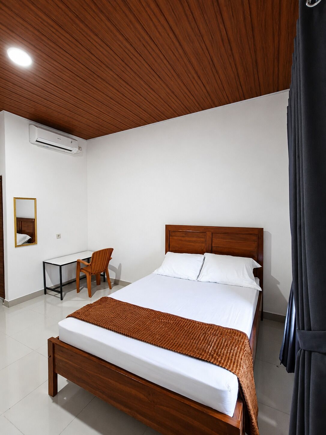 A hotel room with a wooden ceiling, a bed with white bedding, a desk with a chair, and an air conditioning unit.