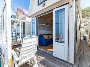 Una terraza de madera con mobiliario exterior, puerta corredera de vidrio que da a una habitación con un sofá azul y una mesita.