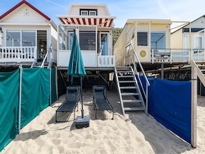 Una propiedad frente a la playa con un área de estar cubierta al aire libre, una escalera que lleva a una terraza y suelo arenoso.