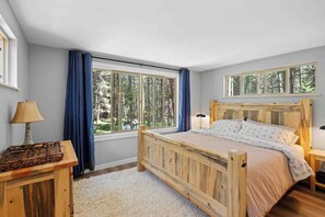 Ein Schlafzimmer mit einem hölzernen Bett, einem Nachttisch, einer Lampe und großen Fenstern mit Vorhängen.