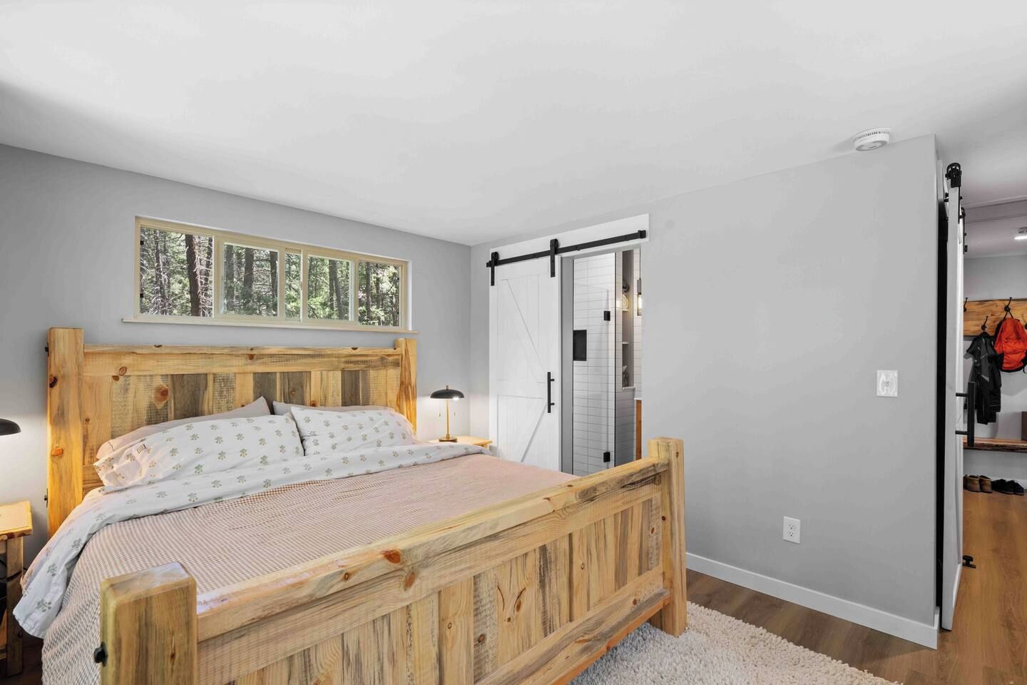 A bedroom with a wooden bed, a window with a forest view, a sliding door, and a coat rack with clothes.