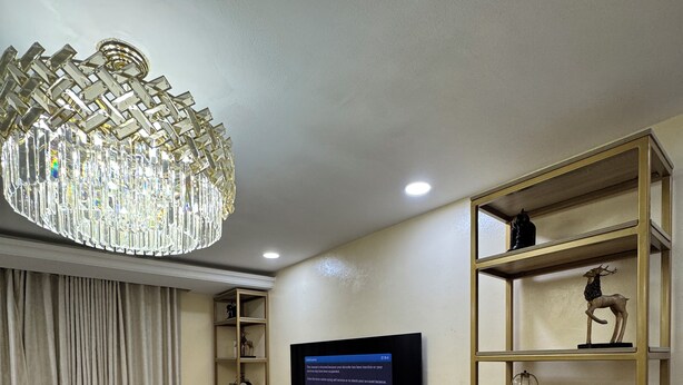 A modern living room with a flat-screen TV mounted on the wall, a wooden console table, a white sofa, and a decorative chandelier. - Spacious 4-bedroom townhome in peaceful Lagos with AC, pool and gym (Lagos)