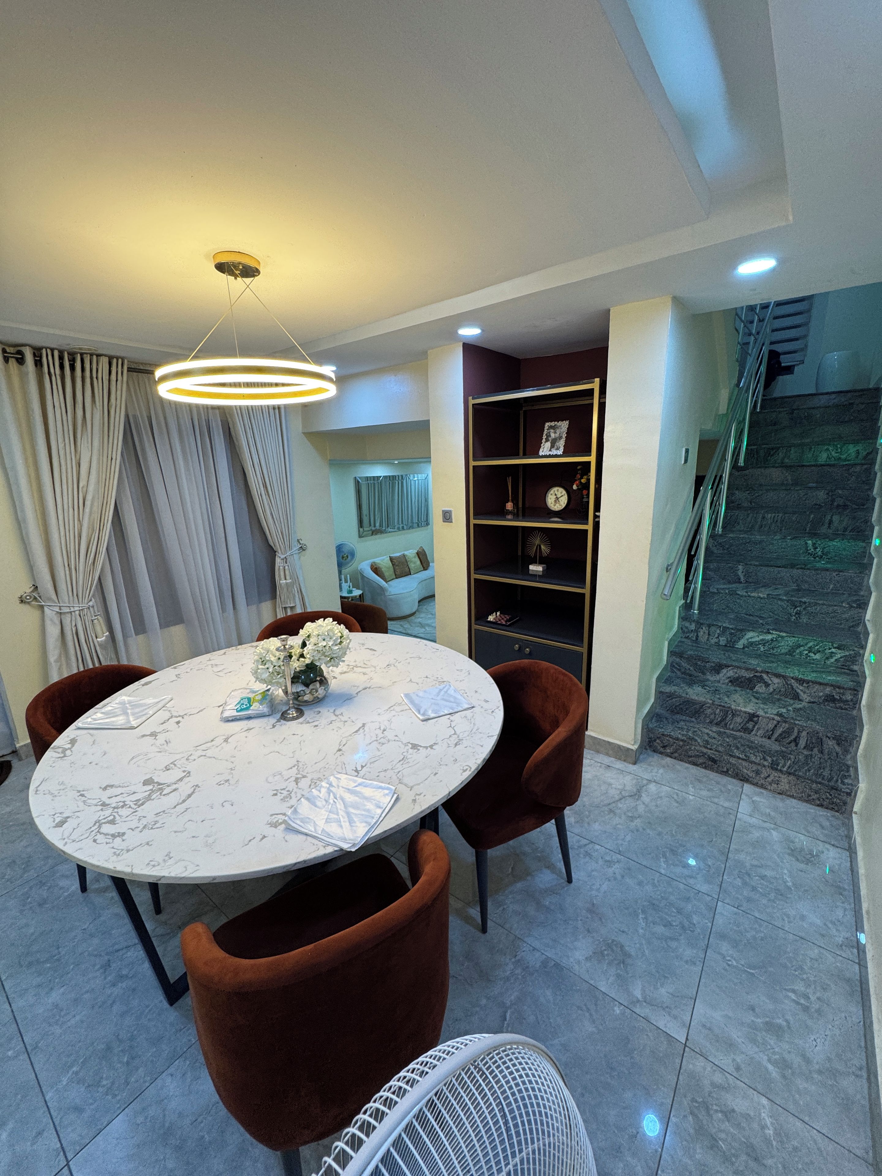 A dining area with a round table, brown chairs, a white fan, and a staircase with a green-lit railing.