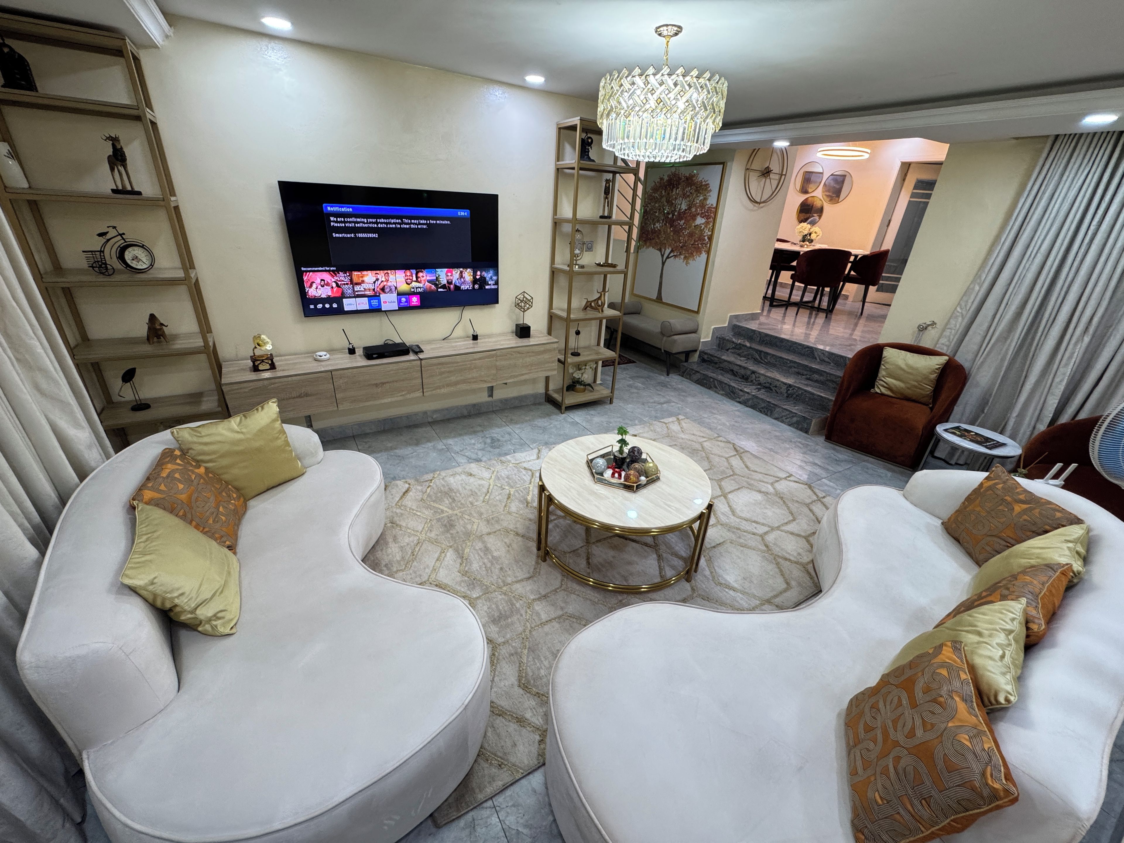 A modern living room with a flat-screen TV, a chandelier, and a staircase leading to a dining area.