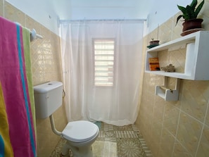 A bathroom with a toilet, a shower curtain, a window with blinds, and a shelf with a plant and a book. - Watamu city comfort apartment, with sea view from rooftop, atm , gym nearby (Watamu)