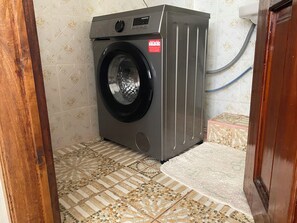 Une petite machine à laver à chargement frontal, de couleur grise, installée dans un petit espace buanderie aux carreaux décoratifs et dotée d’un cadre de porte en bois.