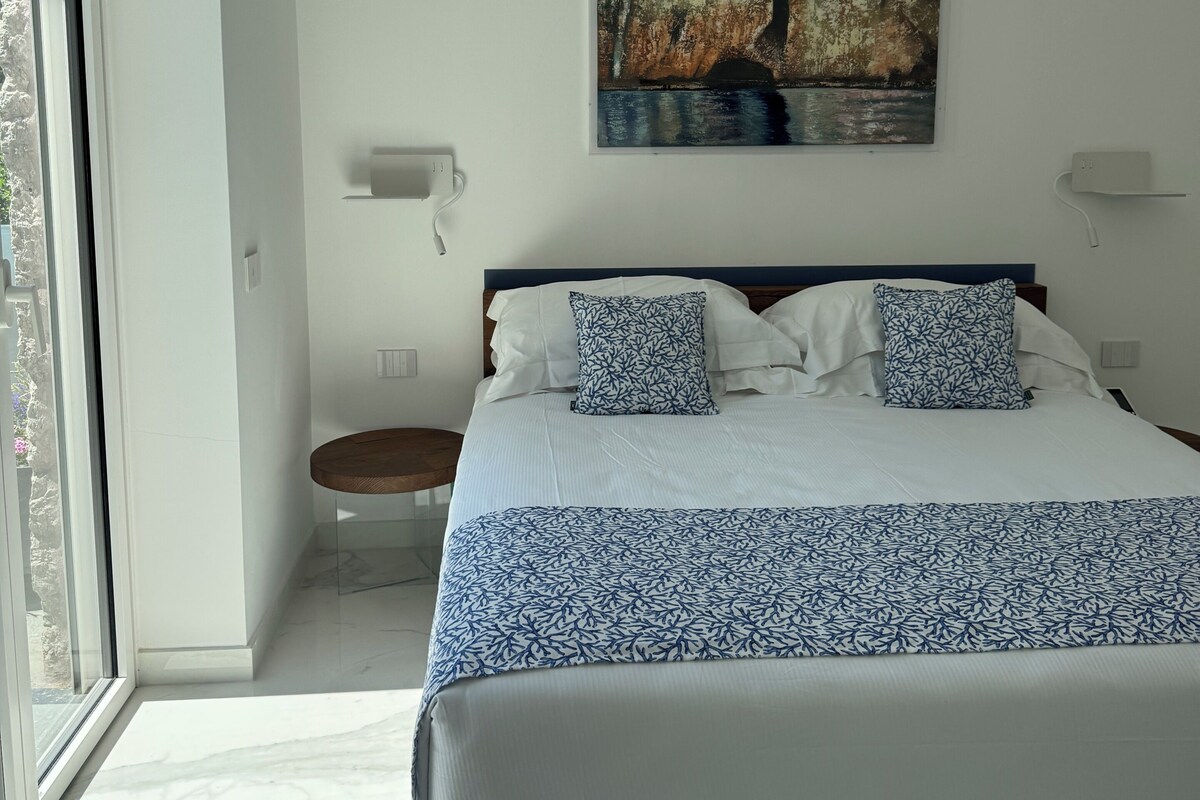 A bedroom with a bed, a painting, a bedside table, and a view of the outdoors.