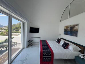 A hotel room with a bed, a TV, a painting, and a view of a coastal area. - Villa L'Ara Due (Capri)