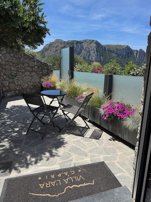 A patio with a table and chairs, a stone wall, and a view of mountains. - Villa L'Ara Due (Capri)