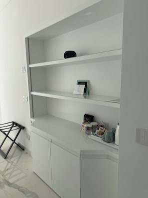 A white cabinet with multiple shelves, a small black object on the top shelf, and a few items on the bottom shelf including a box, packets, and a kettle.
