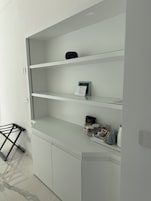 Un mueble blanco con varios estantes, un objeto negro pequeño en el estante superior y algunos artículos en el estante inferior, incluyendo una caja, paquetes y una tetera.