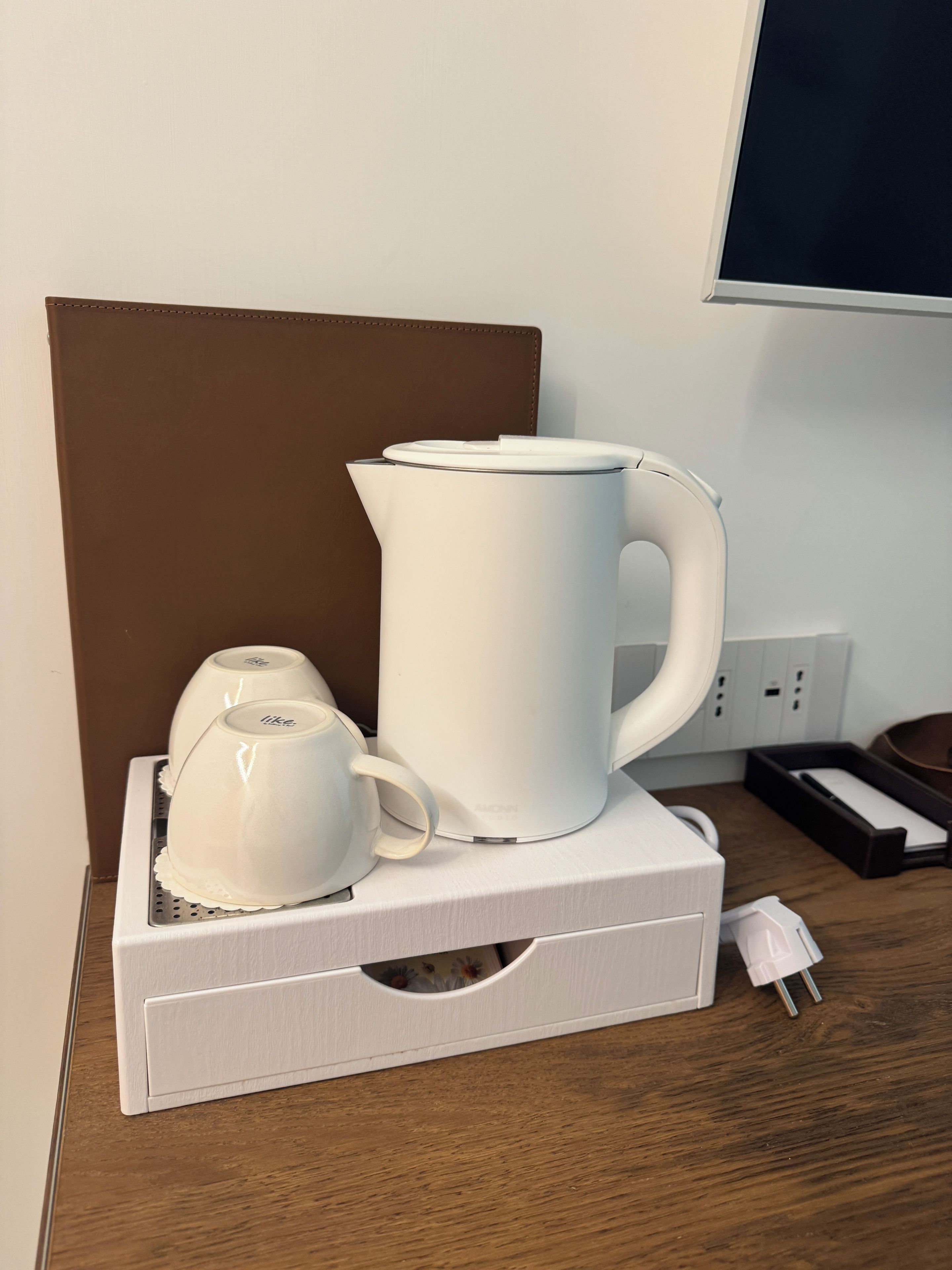 A white kettle and a white mug on a white tray, placed on a wooden table with a brown headboard in the background.
