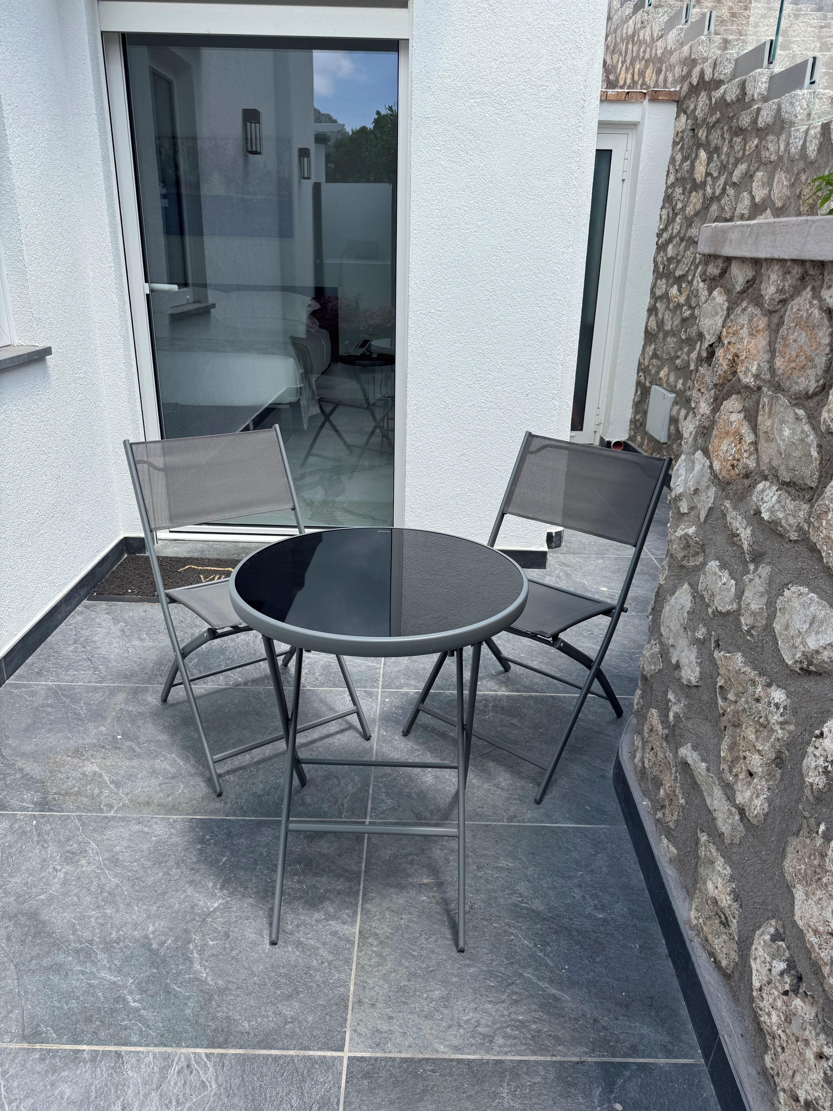 Un patio con una mesa redonda y dos sillas, un muro de piedra y un ventanal de vidrio que da a un espacio interior.