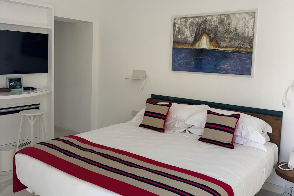 A hotel room with a bed, a desk with a flat-screen TV, a painting on the wall, and a telephone on the nightstand.