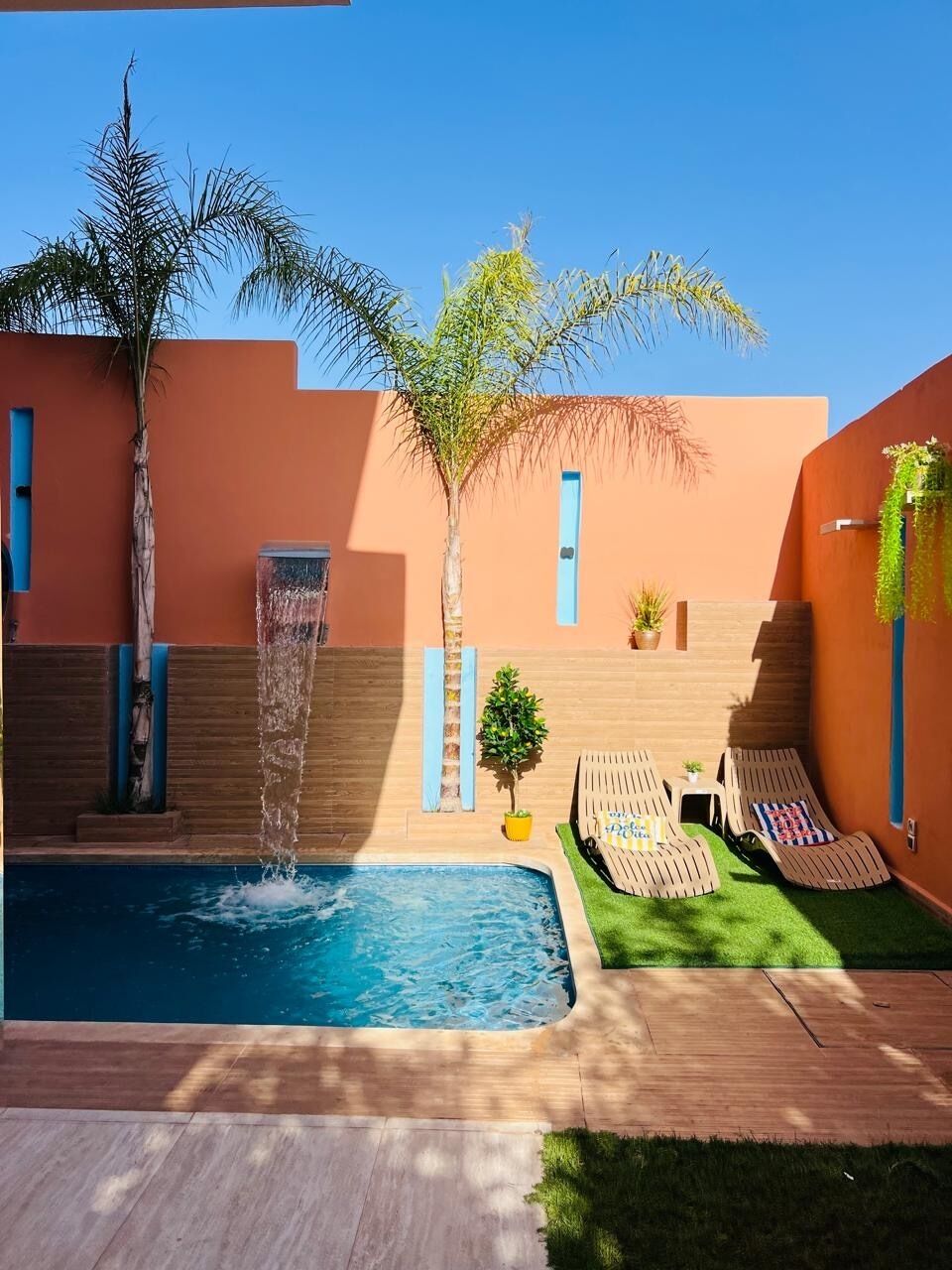 Una piscina amb una font, gandules i una petita zona de jardí amb una palmera i plantes en testos.