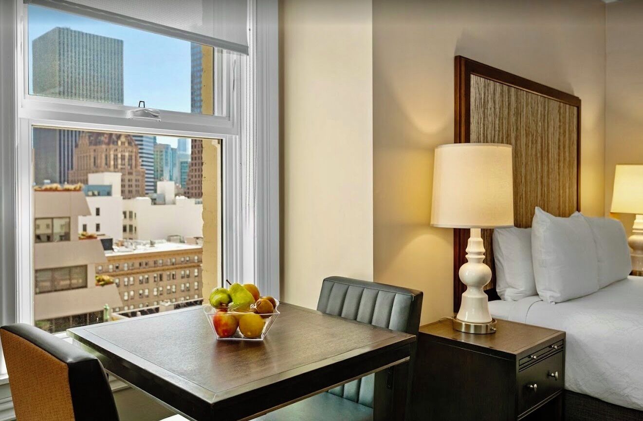 Une chambre d’hôtel avec un lit, une table de chevet avec une lampe, une table à manger avec des fruits, et une vue sur les bâtiments de la ville.