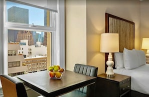 A hotel room with a bed, bedside table with a lamp, a dining table with fruit, and a view of city buildings.