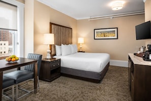 Een hotelkamer met een bed, nachtkastjes, een bureau met stoel, een schaal fruit en uitzicht op de stad.