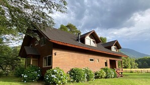 Ein zweigeschossiges Holzhaus mit geneigtem Dach, einer Veranda und einem umzäunten Garten.