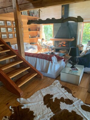 A cozy cabin interior with a wooden staircase, a fireplace, a comfortable seating area, and a bookshelf. - Vrbo Property (Pucon)
