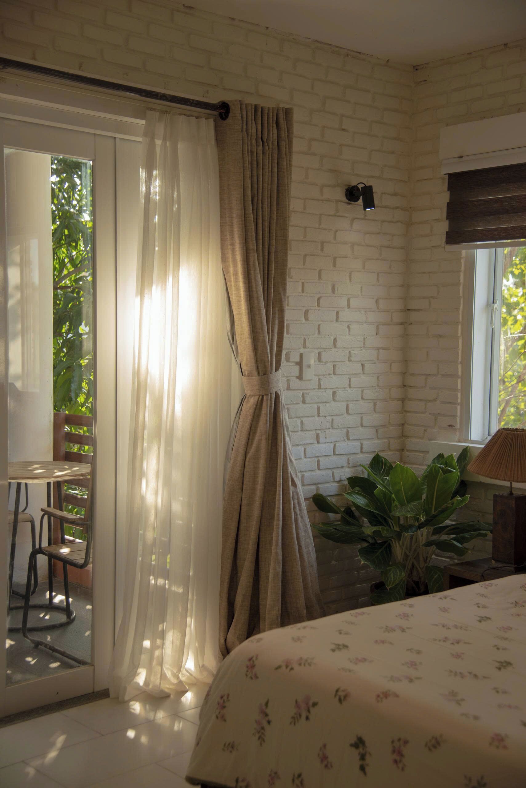 A bedroom with a bed, a window with curtains, a plant, and a small table with chairs.