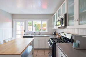 A modern kitchen with white cabinetry, a wooden dining table, and a view of the outdoors through large windows. - Balboa park Craftsman home (San Diego)
